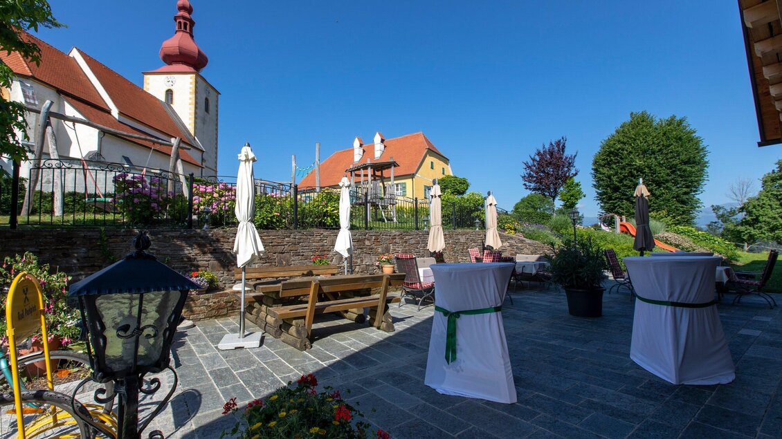 Eine Terrasse mit Tischen und Stühlen, umgeben von Blumen und grünen Bäumen. Im Hintergrund sieht man ein historisches Gebäude mit einem roten Turm und klar blauem Himmel. | © TV Region Graz - Lunghammer