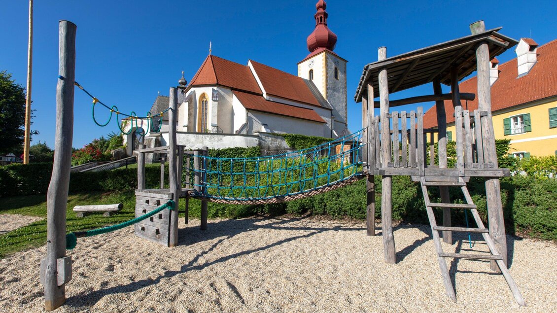 Ein Spielplatz mit einer Spielstruktur aus Holz und einem Klettergerüst. Im Hintergrund ist eine Kirche mit rotem Dach und blauem Himmel zu sehen. | © TV Region Graz-Lunghammer