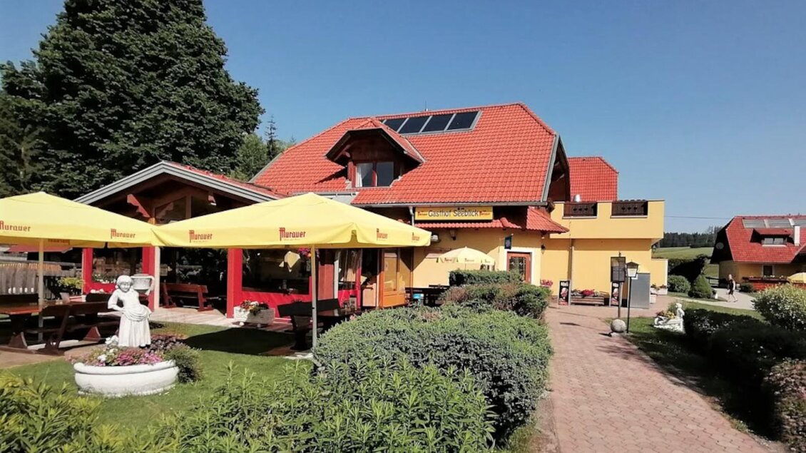 Ein Restaurant mit einer gelben Fassade und einem roten Dach. Draußen stehen Terrassen mit Sonnenschirmen und eine grüne Gartenlandschaft. | © Gasthof Seeblick