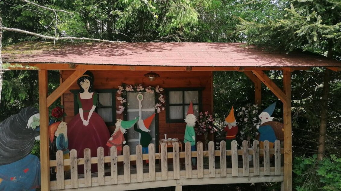 Ein kleines Holzhaus mit einer bunten Wandmalerei von Märchenfiguren, darunter eine Prinzessin und Zwerge. Der Bereich ist von einem weißen Zaun umgeben und von Bäumen umgeben. | © Gasthof Seeblick