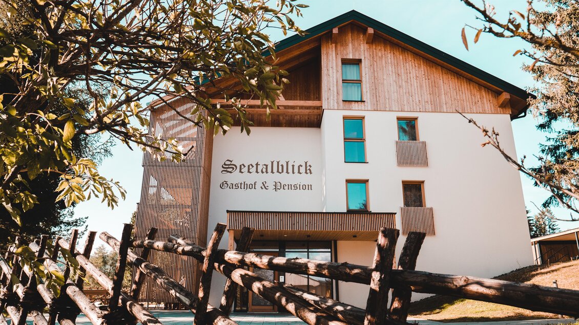 Ein modernes Gasthaus mit Holzverkleidung und einem einladenden Eingang. Umgeben von Bäumen und einem Zaun wirkt es freundlich und einladend. | © Gasthof und Pension Seetalbllick