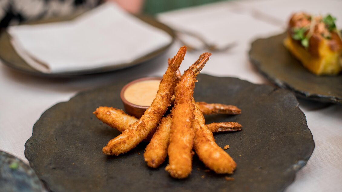 Frittierte Garnelen auf einem schwarzen Teller, dazu eine kleine Schale mit Dipsauce. Im Hintergrund sind Servietten und andere Speisen zu sehen. | © Erlebnisregion Murtal