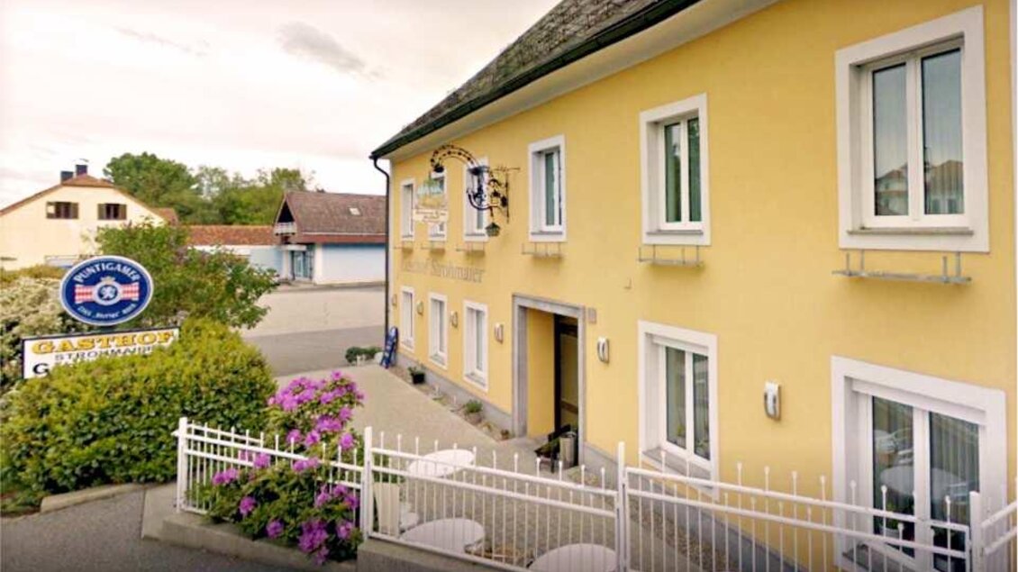 Ein gelbes Gebäude mit weißen Fenstern und einem schönen Garten. Im Vordergrund befindet sich ein Zaun mit Blumen. | © Strohmeier