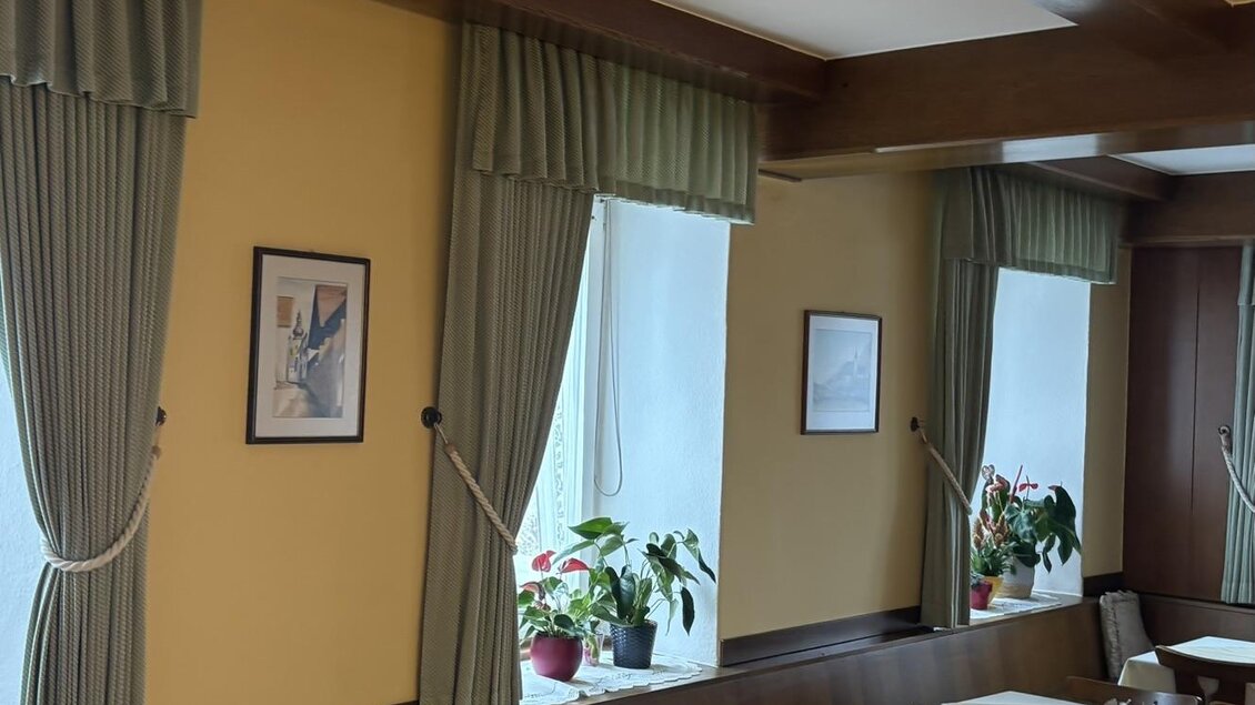 Ein gemütlicher Innenraum mit hellen Tischen und Holzstühlen. An den Fenstern hängen grüne Vorhänge und es stehen Pflanzen auf den Fensterbänken. | © Strohmeier
