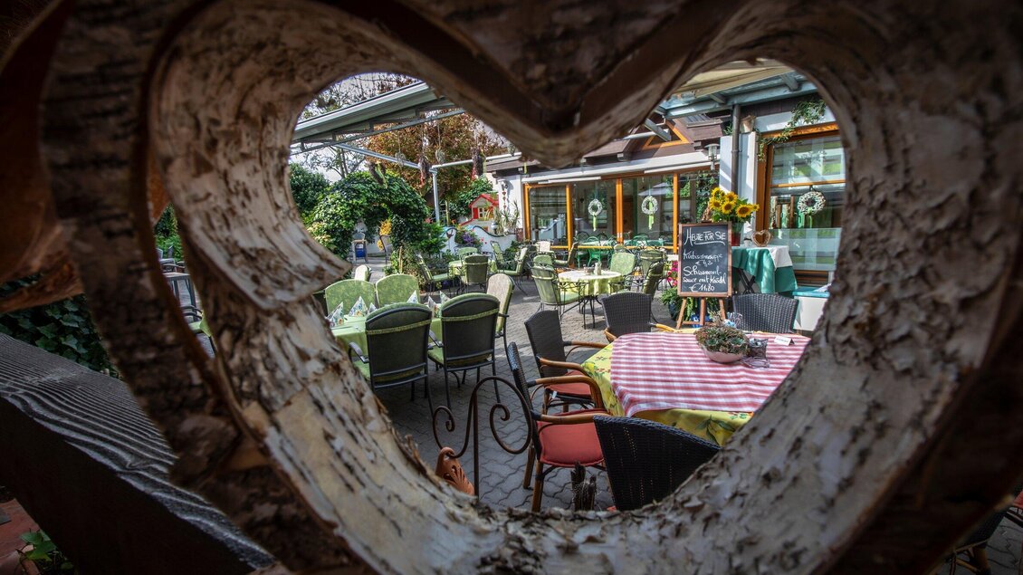 Ein gemütlicher Restaurantbereich mit Tischen und Stühlen im Freien. Der Blick ist durch eine herzförmige Öffnung gerahmt. | © TV Region Graz - Lunghammer
