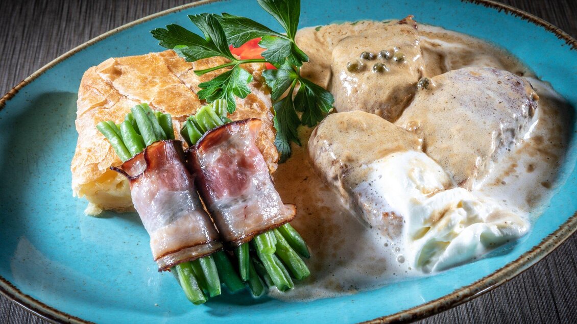 Ein köstliches Gericht mit zartem Fleisch in einer cremigen Soße, umgeben von grünen Bohnen und einer Blätterteigbeilage. Ein dekoratives Element aus Petersilie rundet das ansprechende Essen ab. | © TV Region Graz - Lunghammer