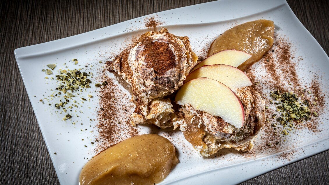 Ein Teller mit Dessert, bestehend aus zwei Portionen cremiger Masse, dekoriert mit Apfelscheiben und Zimt. Dazu gibt es eine Portion Apfelmus und zerstoßene Pistazien. | © TV Region Graz - Lunghammer