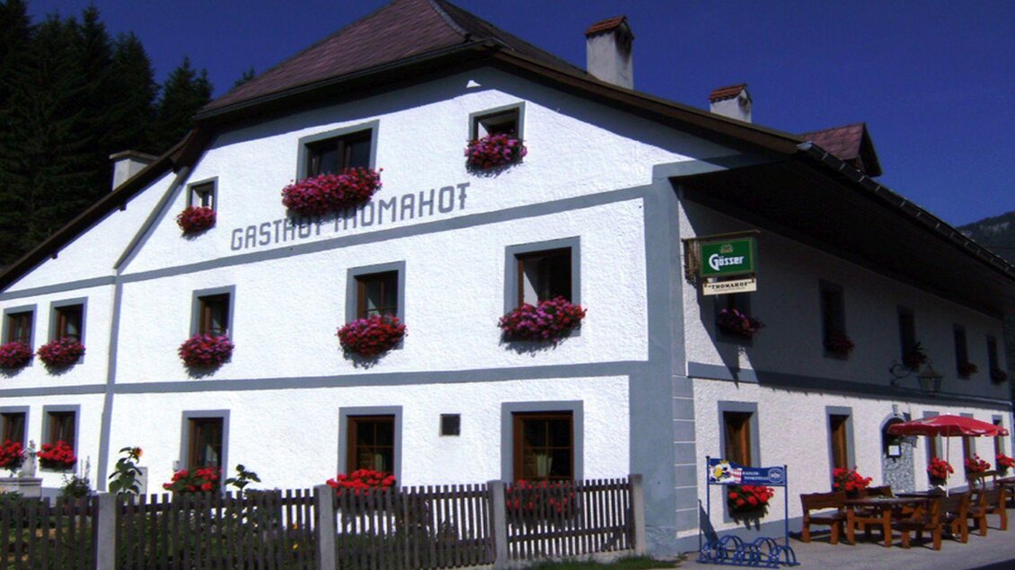 Ein traditionelles Gasthaus mit weißer Fassade und bunten Blumenkästen. Draußen stehen Tisch und Stühle für Gäste. | © Gasthof Thomahof