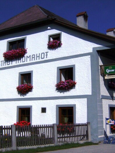 A traditional inn with a white facade and colorful flower boxes. Outside, there are tables and chairs for guests. | © Gasthof Thomahof