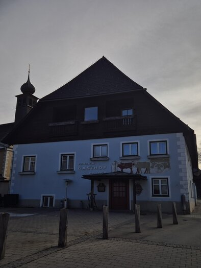 A large blue building with a pointed roof and a church tower in the background. The surroundings are quiet, with a few trees and an asphalted road. | © Tourismusverband Murau