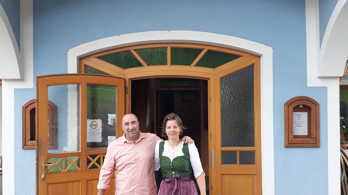 Ein Mann und eine Frau stehen vor dem Gasthof Wiendl. Die Frau trägt ein traditionelles Bauernkleid, während der Mann lässige Kleidung trägt. | © TV Lipizzanerheimat