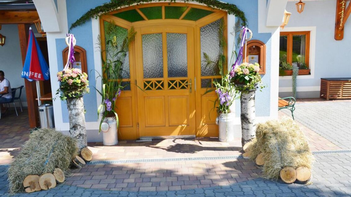 Eine einladende Eingangstür mit bunten Blumenarrangements und dekorativem Heu. Die Fassade ist hellblau und strahlt eine freundliche Atmosphäre aus. | © Gasthof Wiendl