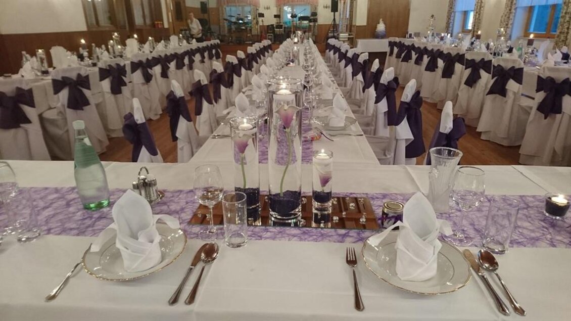 Ein festlich dekorierter Saal mit langen Tischen, auf denen Tischdecken und Servietten in Weiß und Lila liegen. In der Mitte der Tische stehen dekorative Gläser und Kerzen für eine elegante Atmosphäre. | © Gasthof Wiendl
