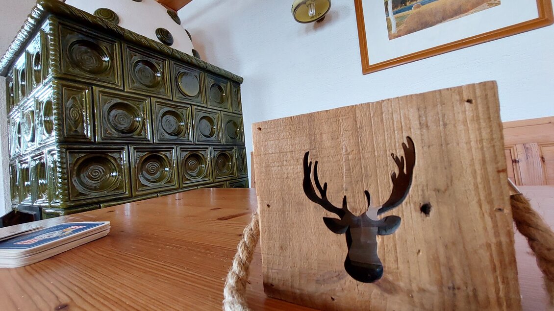 Ein rustikaler Tisch aus Holz mit einem dekorativen Holzschild, das ein Geweih zeigt. Im Hintergrund steht ein traditioneller keramikfarbener Ofen. | © TV Hochsteiermark / Brigitte Digruber
