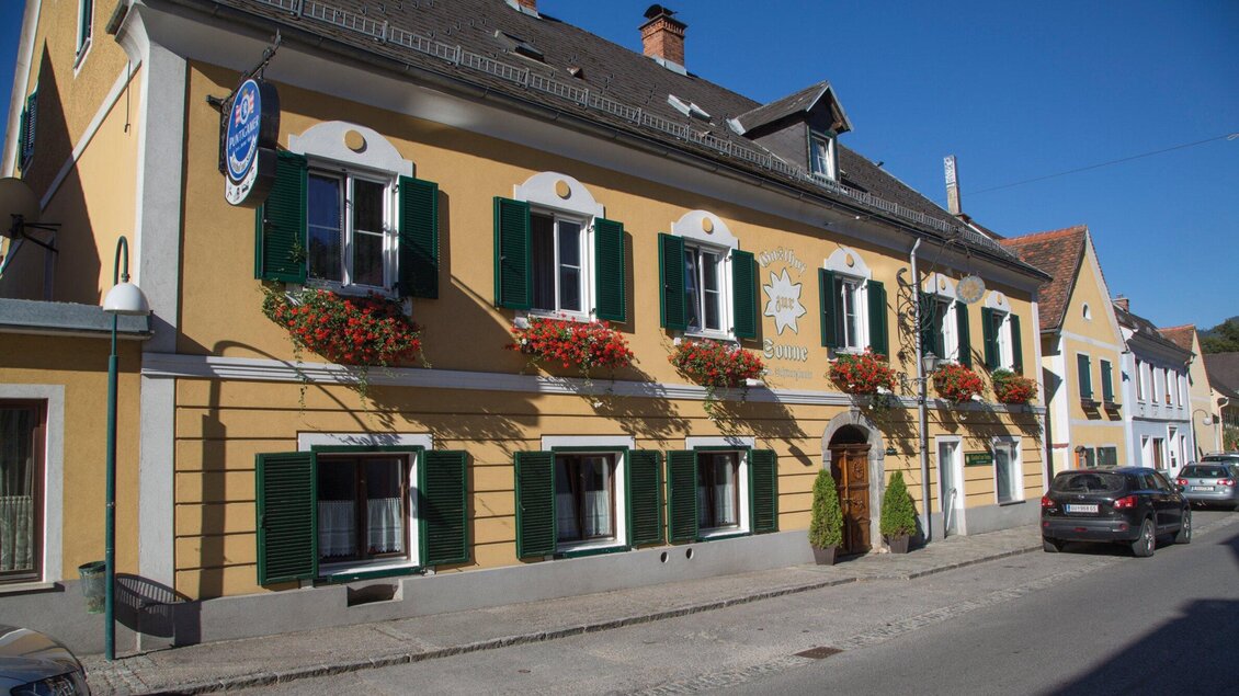 Ein charmantes gelbes Gebäude mit grünen Fensterläden und Blumenkästen. Die Straße vorne ist ruhig und typisch für eine malerische Stadt. | © TV Region Graz - Schwarzbauer