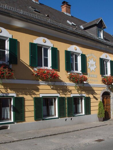 A charming yellow building with green shutters and flower boxes. The street in front is quiet and typical for a picturesque town. | © TV Region Graz - Schwarzbauer