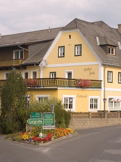 Ein gelbes Hotel mit mehreren Fenstern und einem schönen Balkon. Es liegt an einer Straßenecke und ist von Bäumen und Blumen umgeben. | © Gasthof Jaritz - Kreuzwirt