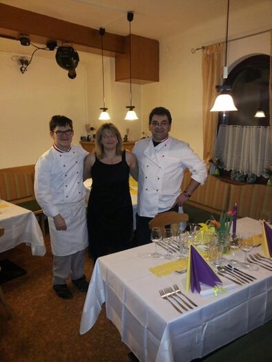 An elegant restaurant with set tables and a warm atmosphere. Three people, including uniformed chefs, are posing happily for the photo. | © Gaststätte Franz August