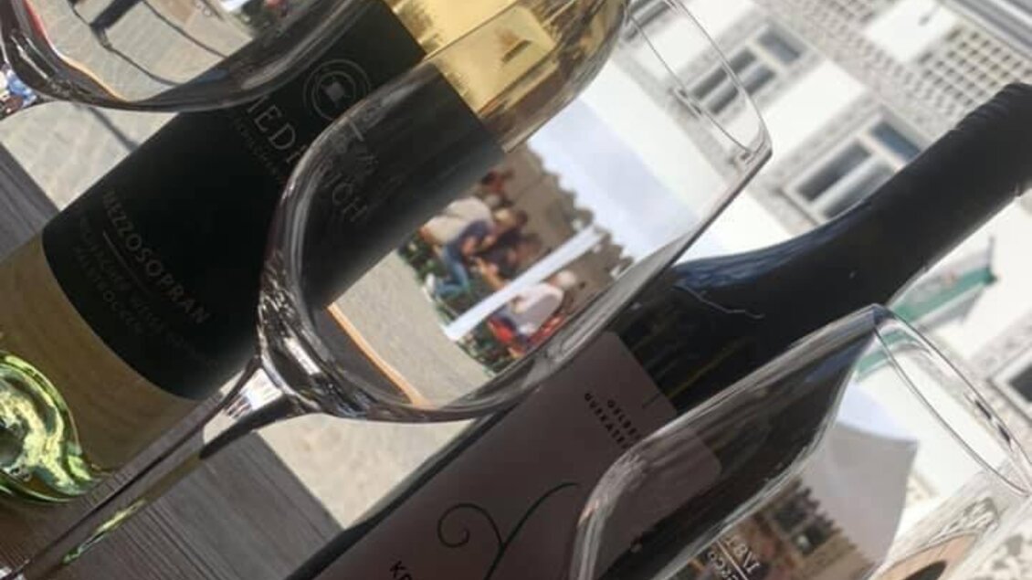 Zwei Weinflaschen stehen vor zwei Weingläsern auf einem Tisch. Im Hintergrund sind Menschen und eine malerische Stadtansicht zu sehen. | © Gaststätte Franz August