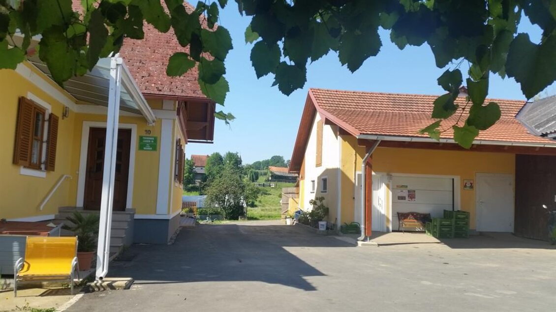 Ein ruhiger Hof mit zwei gelben Häusern und blauen Himmel. Im Hintergrund sind grüne Wiesen und Bäume sichtbar. | © Tschernko