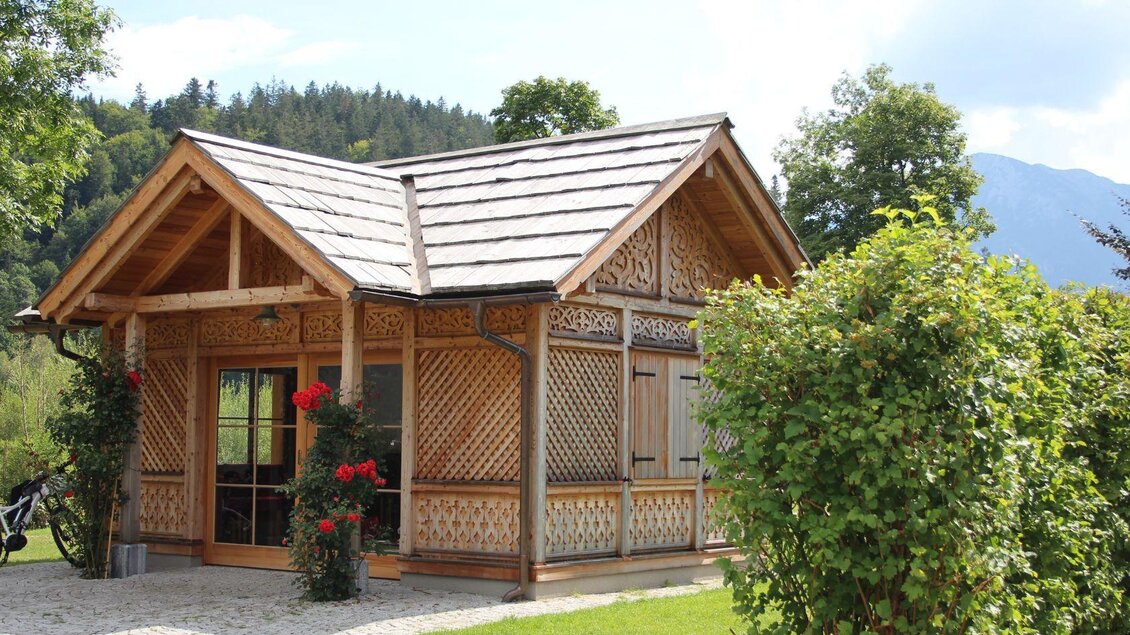 Ein charmantes Holzhaus mit kunstvollen Verzierungen und einem gepflegten Garten. Umgeben von Bäumen und sanften Hügeln bietet es eine idyllische Aussicht. | © Viola Lechner