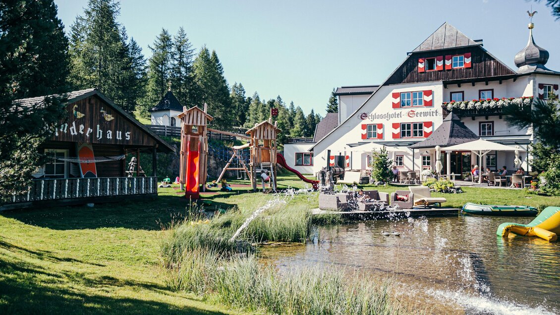 Ein malerisches Gelände mit zwei Gebäuden, Spielplatz und einem kleinen Teich. Umgeben von grünen Bäumen und einem einladenden Garten. | © Schlosshotel Seewirt