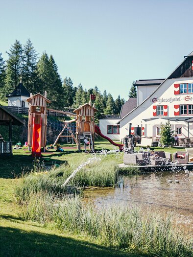 A picturesque area with two buildings, a playground, and a small pond. Surrounded by green trees and an inviting garden. | © Schlosshotel Seewirt