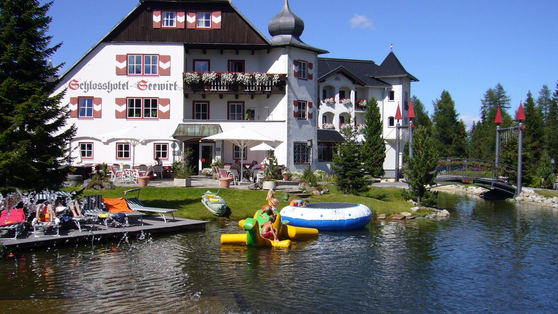 Ein malerisches Hotel am Wasser mit einer grünen Umgebung. Im Vordergrund genießen Personen die Freizeitaktivitäten auf einem Spielboot. | © Schlosshotel Seewirt