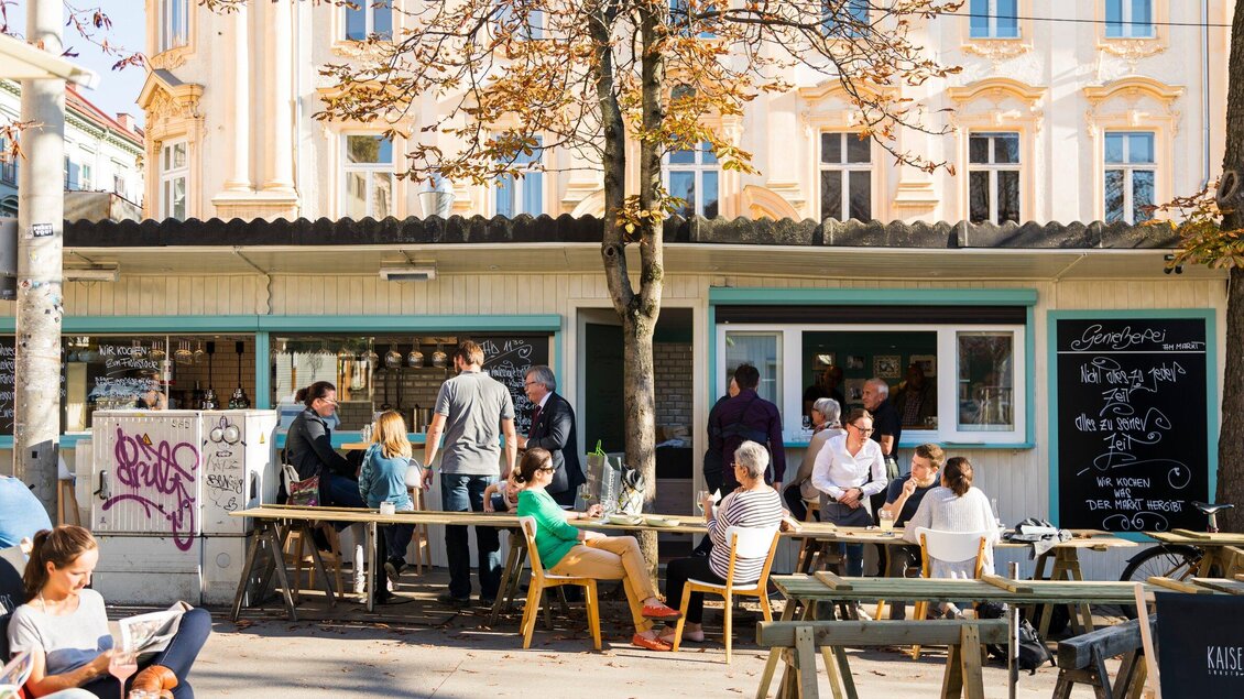 Ein belebtes Café mit Gästen, die draußen an Holztischen sitzen. Im Hintergrund sind niedrige Gebäude und ein Baum zu sehen. | © Genießerei am Markt