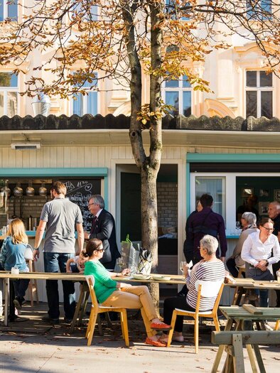 A bustling café with guests sitting outside at wooden tables. In the background, there are low buildings and a tree visible. | © Genießerei am Markt