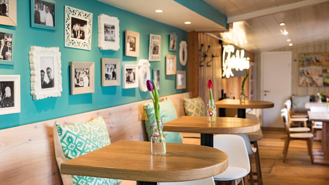 Ein gemütliches Café mit Holztischen und einer blauen Wand dekoriert mit Bildern in verschiedenen Rahmen. Auf den Tischen stehen Vasen mit frischen Blumen. | © Genießerei am Markt - Werner Krug