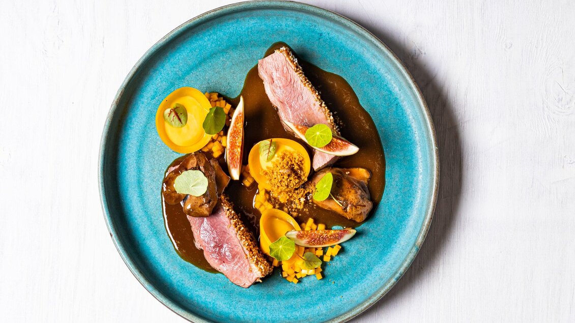 Ein kunstvoll angerichteter Teller mit gebratenem Fleisch, frischen Feigen und orangefarbenen Früchten. Die bunte Präsentation wird durch eine dunkle Soße und gehackte Zutaten ergänzt. | © Genießerei am Markt