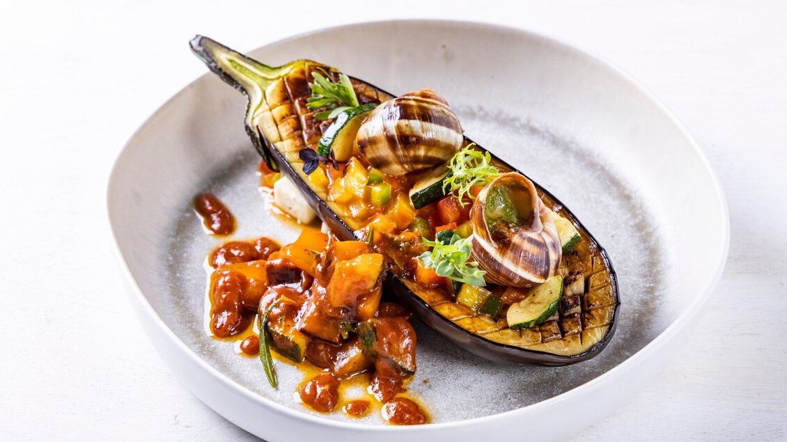 Ein gegrilltes Auberginengericht in einer weißen Schüssel. Es ist mit buntem Gemüse und einer Sauce garniert. | © Genießerei am Markt - Werner Krug