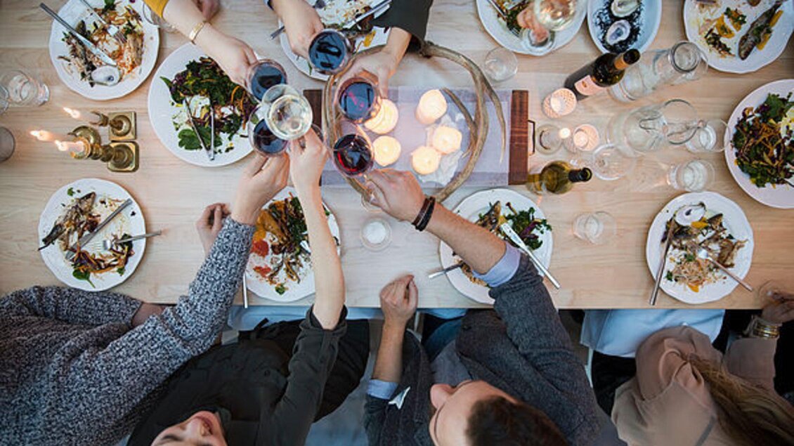 Ein Tisch, der mit verschiedenen Gerichten und Getränken gedeckt ist. Menschen stoßen mit Gläsern an und genießen eine gesellige Mahlzeit. | © Genießerei am Markt