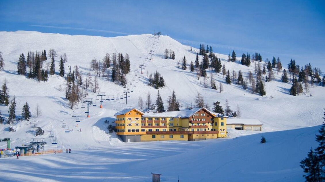 Ein schneesicheres Bergresort mit einem gelben Hotel. Im Hintergrund sind verschneite Pisten und hohe Bäume zu sehen.