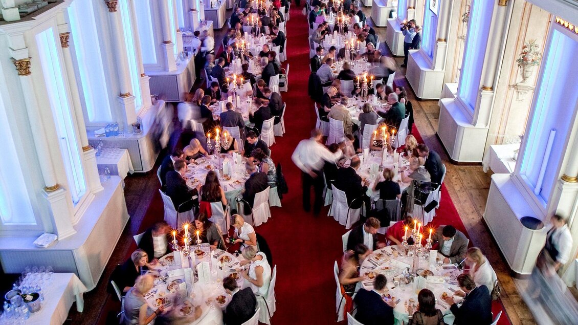 Ein elegantes Bankett mit festlich gedeckten Tischen und Kerzenlicht. Viele Gäste genießen das Abendessen in einem stilvollen Raum mit blauen Lichtern. | © Edlers