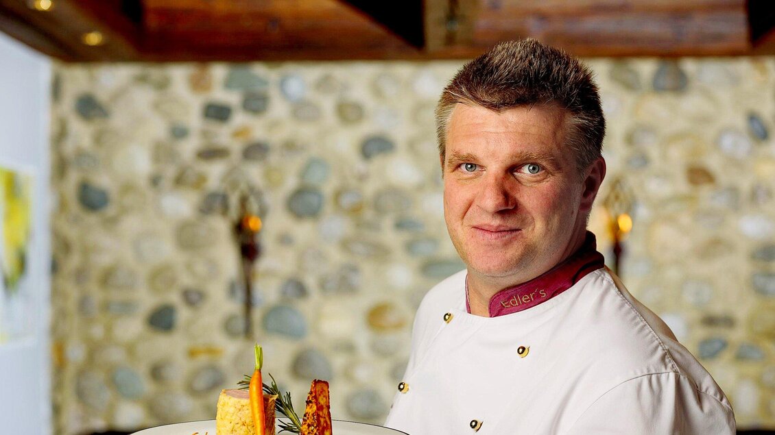 Ein Koch präsentiert stolz ein Gericht auf einem Teller. Im Hintergrund ist eine steinerne Wand sichtbar. | © Edlers