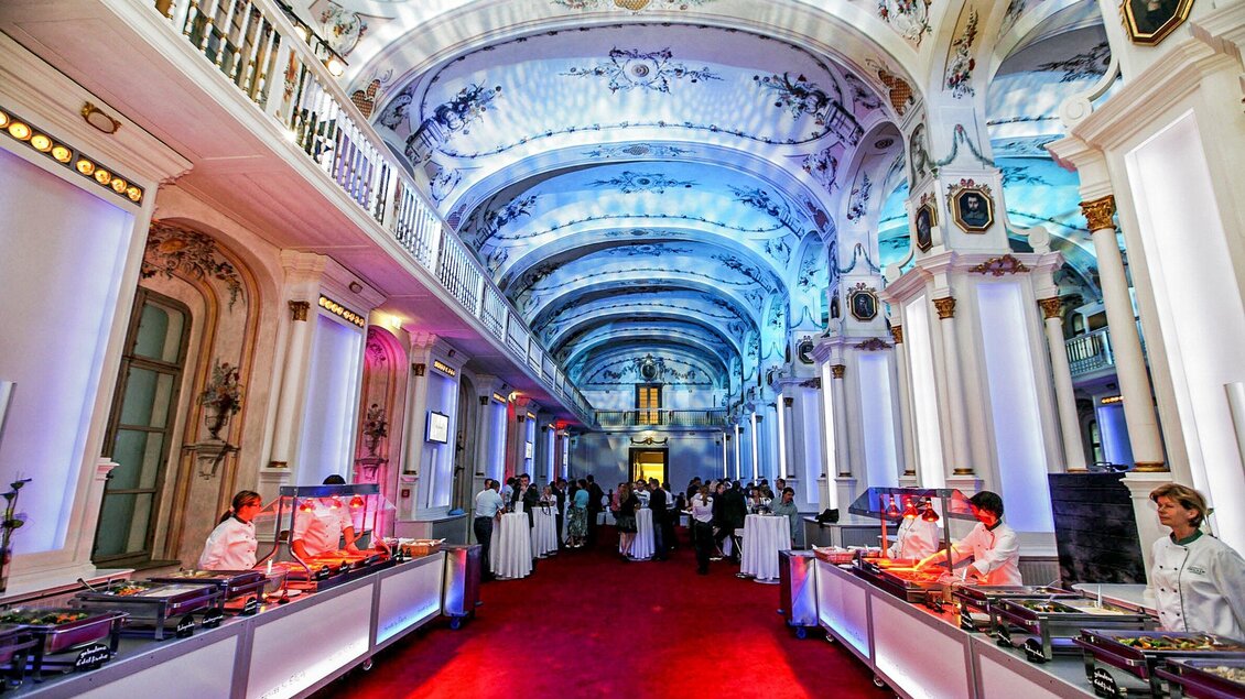 Ein eleganter Saal mit hohen Decken und buntem Licht. In der Mitte des Raumes stehen Tische mit Gästen und Köchen, die Speisen zubereiten. | © Edlers