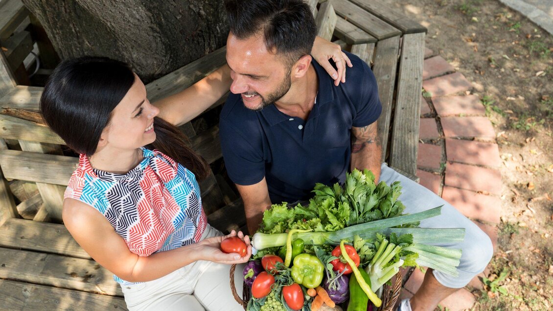 Ein Paar sitzt auf einer Bank und lächelt sich an. Sie halten einen Korb mit frischem Gemüse und Kräutern in der Hand. | © Region Graz - Harry Schiffer