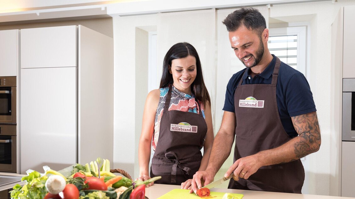 Ein Paar kocht in einer modernen Küche und schneidet frisches Gemüse. Beide tragen Schürzen und lächeln, während sie gemeinsam arbeiten. | © Region Graz - Harry Schiffer