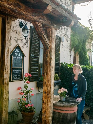 Frau steht an einem Holztisch vor traditioneller Gaststätte im Grünen | © Graz Tourismus - Mias Photoart