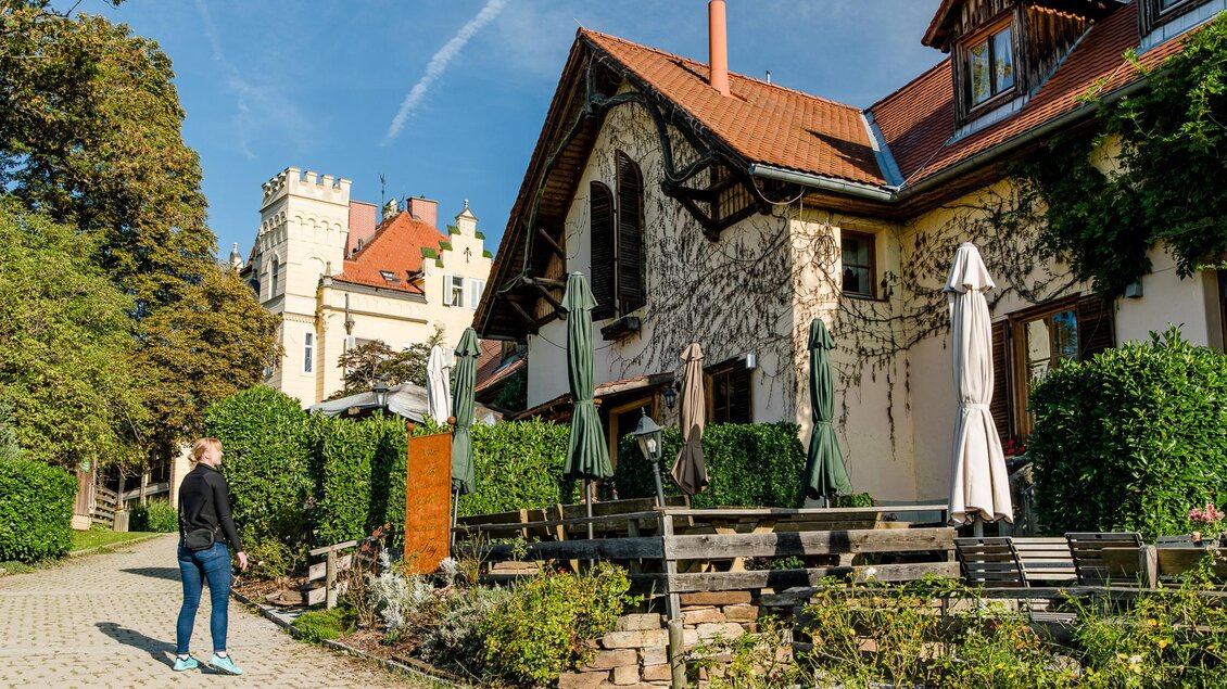 Frau geht an Terrasse mit Sonnenschirmen vorbei, im Hintergrund ein Schloss | © Graz Tourismus - Mias Photoart