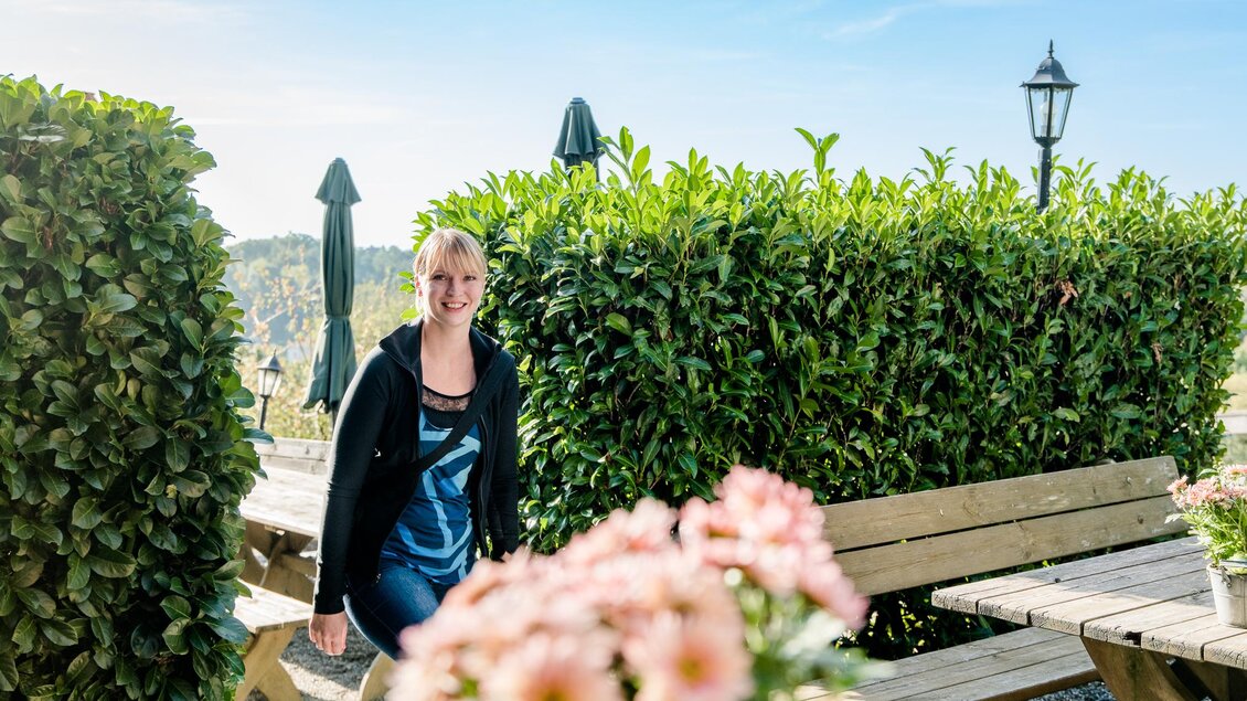 Frau sitzt lächelnd an Holztisch zwischen Hecken und Blumen | © Graz Tourismus - Mias Photoart