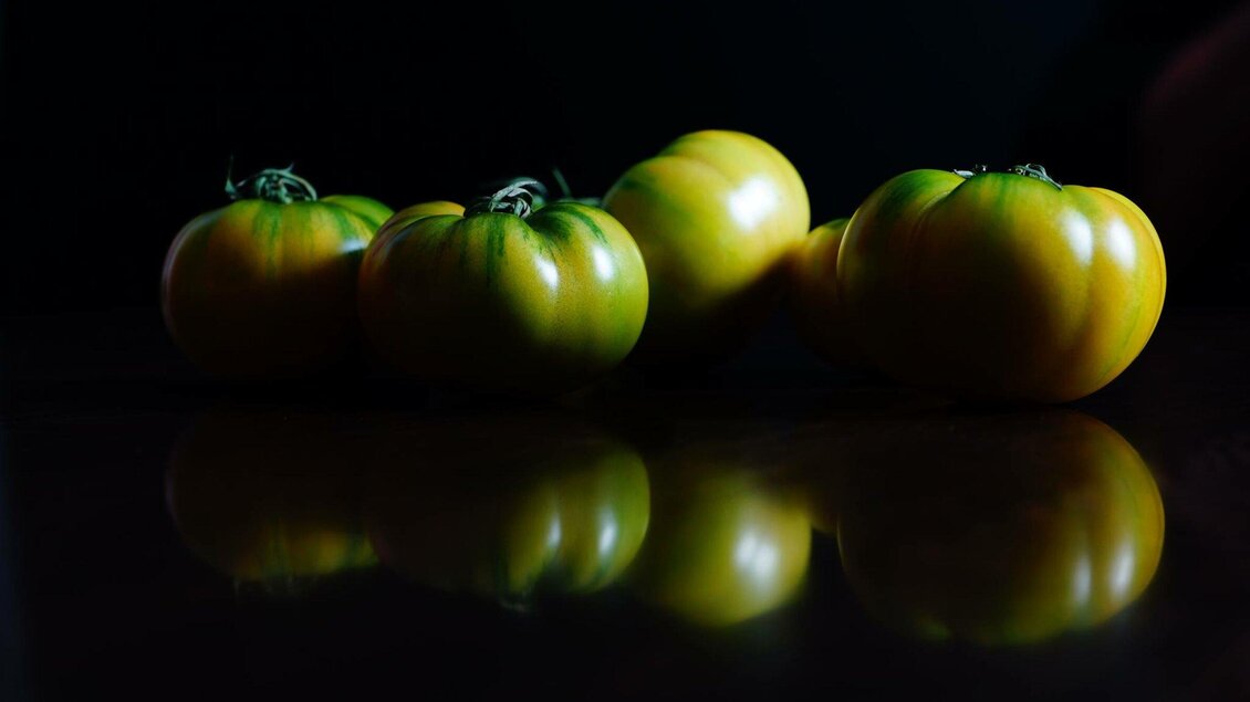 Fünf grüne Tomaten stehen auf einer dunklen Oberfläche. Ihr Glanz spiegelt sich im Untergrund wider. | © Gerüchteküche