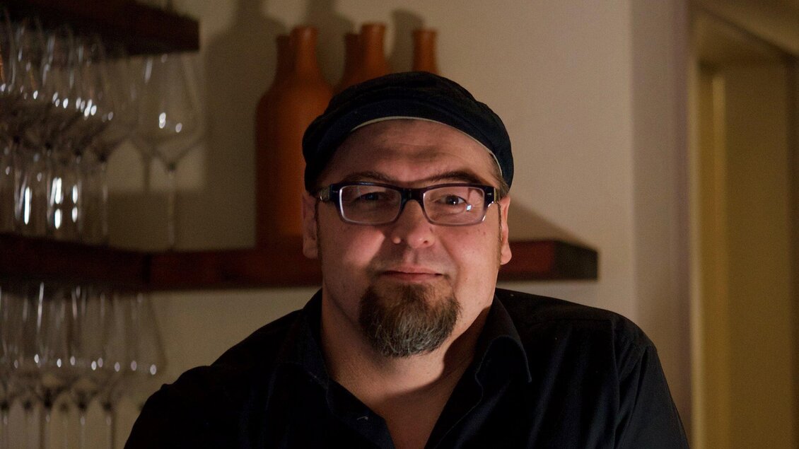 Ein Mann mit Brille und Hut steht in einem gemütlichen Raum. Im Hintergrund sind Weingläser und dekorative Vasen zu sehen. | © Gerüchteküche