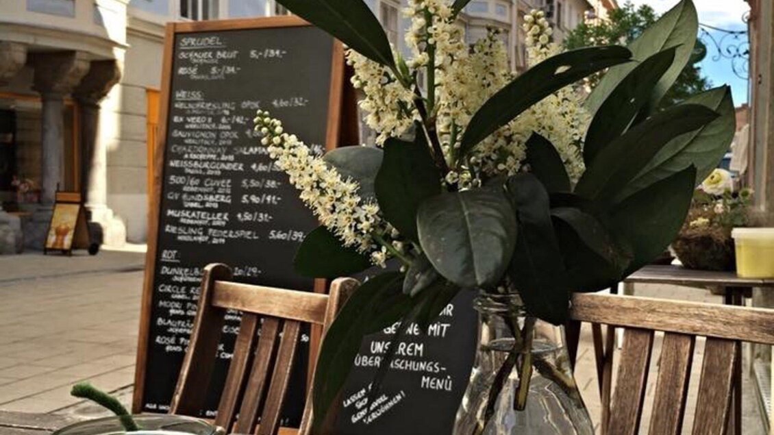 <p>Ein Tisch im Freien mit einer Vase mit Blumen und Blättern. Im Hintergrund ist eine Tafel mit Menüpunkten zu sehen.</p> | © Gerüchteküche