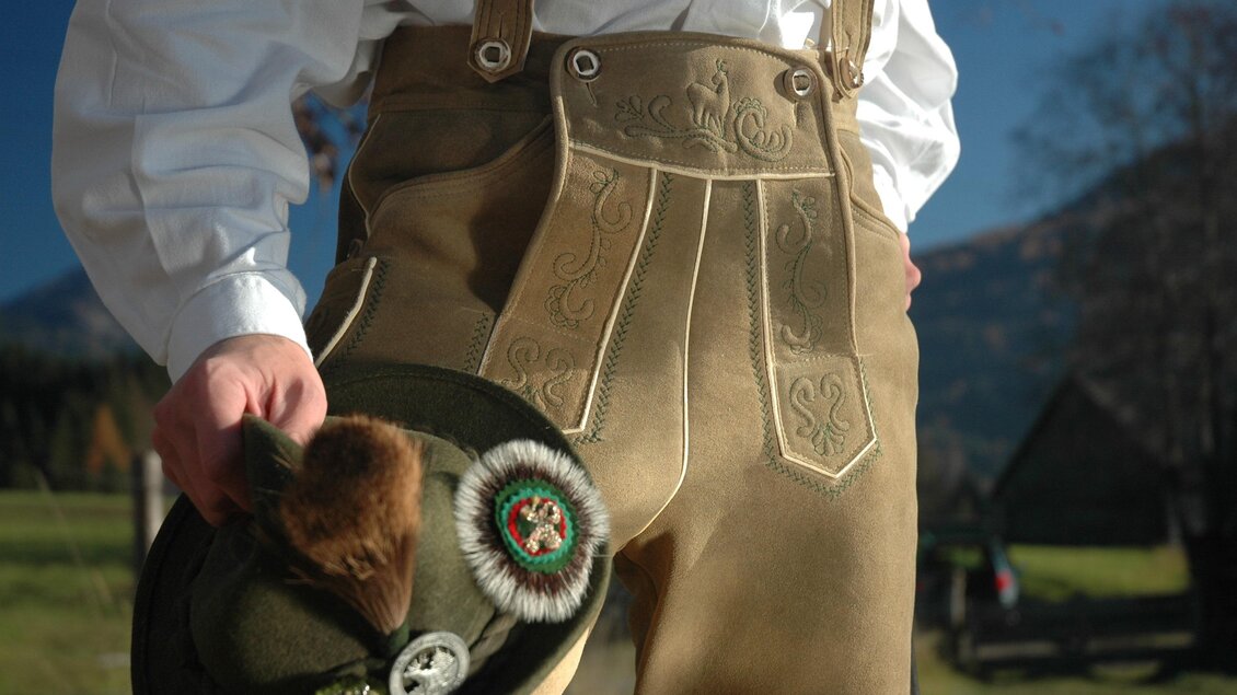 Ein traditionelles Outfit mit Lederhosen und einem Hut. Die Umgebung wirkt ländlich und sonnig. | © Gerberei Schlüßlmayr 