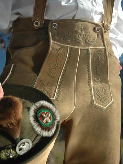 Ein traditionelles Outfit mit Lederhosen und einem Hut. Die Umgebung wirkt ländlich und sonnig. | © Gerberei Schlüßlmayr 