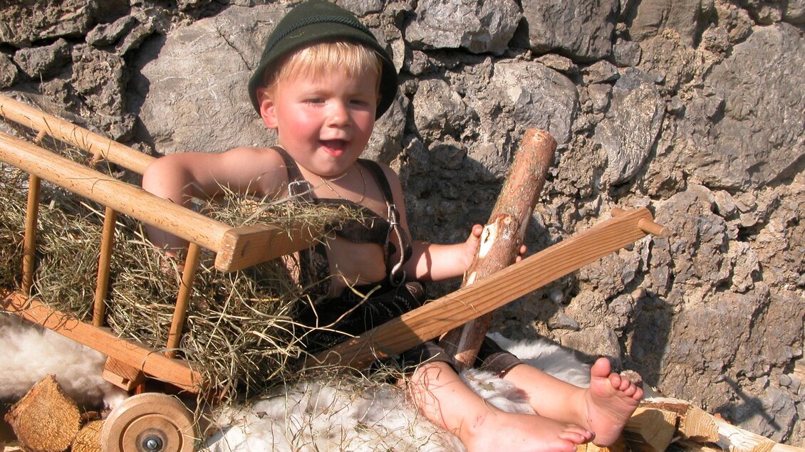 Ein kleiner Junge sitzt in einer Holzschubkarre voller Heu. Er trägt einen Hut und sieht fröhlich aus, während er auf einem Holzstapel sitzt. | © Gerberei Schlüßlmayr 