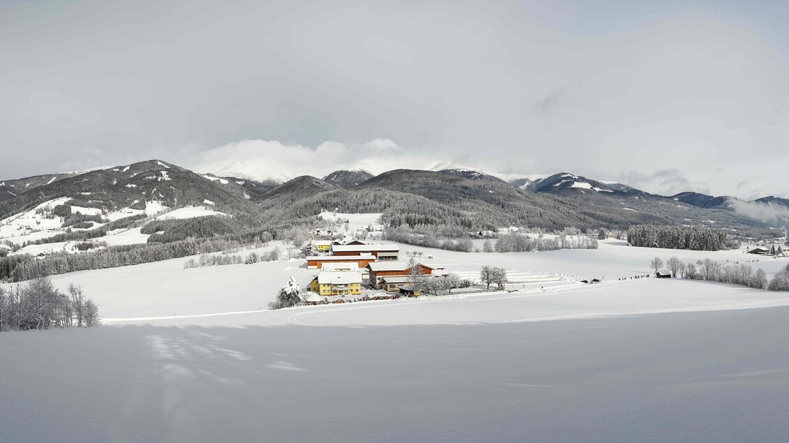 Eine winterliche Landschaft mit einer schneebedeckten Ebene und sanften Hügeln. In der Ferne ist ein Bauernhof zu sehen, umgeben von schneebedeckten Bäumen und Bergen. | © Fam. Offenbacher